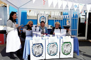 St Kilda Mums fundraising stall featuring volunteers with custom printed organic cotton tea towels and aprons, raising funds for families in need in Australia.