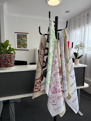 Tea towels displayed on a stand featuring customisable print designs from Tea Towels Australia’s free template collection, including illustrated teapots, native herbs, and Australian motifs.