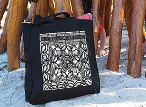 Black cotton tote bag with intricate mandala-style custom print, resting on sand beside wooden play structure
