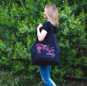 Woman carrying a custom-printed black tote bag with colourful artwork, standing in front of green foliage