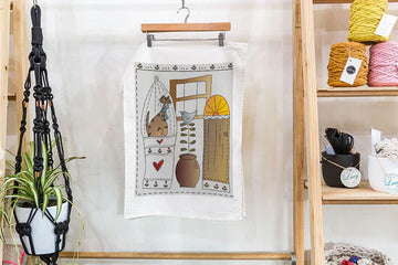 Linen blend tea towel featuring Theodora Cleave’s watercolour artwork of a cat sitting by a window with potted plants, hanging on a wooden rack in a cozy craft studio.