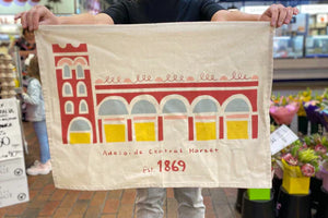 Person holding a custom printed tea towel featuring a stylised illustration of the Adelaide Central Market building with 'Est. 1869' and vibrant red, yellow,blue and pink artwork, designed for Expressions Tea Towels Australia.