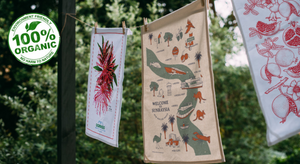 Three 100% organic cotton tea towels with botanical and illustrated designs hanging outdoors against a green leafy background