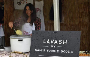 MasterChef’s Samira Damirova serving food at a market stall wearing a custom printed apron, with tea towels displayed behind and a Lavash by Sam’s Foodie Goods sign in front.