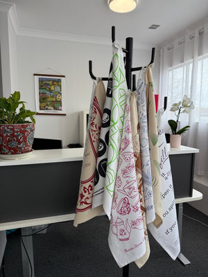 Tea towels displayed on a stand featuring customisable print designs from Tea Towels Australia’s free template collection, including illustrated teapots, native herbs, and Australian motifs.