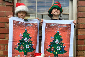 Two children wearing Christmas hats holding personalised Christmas tea towels featuring a decorated tree design from Expressions Tea Towels Australia