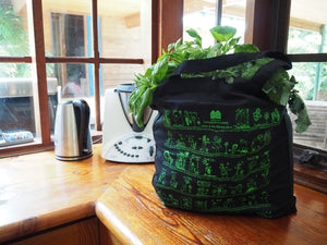 Black organic cotton tote bag printed with children's drawings in green ink, filled with fresh vegetables including basil and broccoli, sitting on a wooden kitchen bench near a Thermomix and kettle.