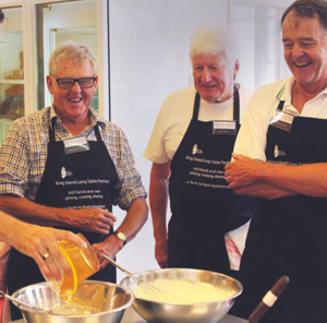 Three men wearing personalised black aprons with white printed logos, smiling and cooking together in a kitchen during a community or team event.