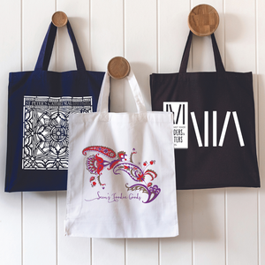 Three custom printed canvas tote bags hanging on wooden hooks against a white panel wall, showcasing different personalised designs and branding styles.