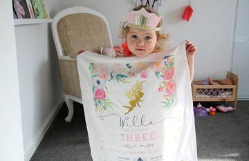 Little girl wearing a pink crown holding a personalised fairy-themed organic cotton tea towel birthday invitation in a playroom