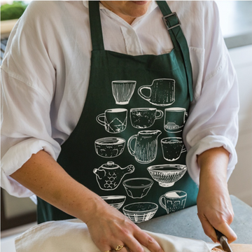 Person wearing a dark green personalised apron with white illustrated coffee cups and bowls, slicing food on a kitchen counter.