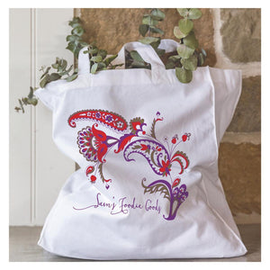 White cotton tote bag with colourful paisley-style floral design and 'Sam's Foodie Goody' text, filled with eucalyptus branches, styled against a stone wall.