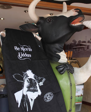 Black personalised apron with humorous ‘Be Nice to Udders’ slogan and cow illustration, displayed on a life-sized cow statue at a dairy-themed venue in Australia