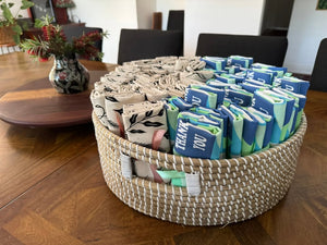 Woven basket filled with neatly folded custom printed tea towels, including blue “Thank You” designs and beige botanical prints, displayed on a wooden table.