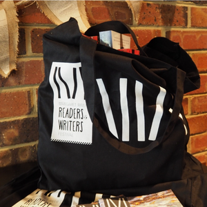 Black custom tote bag with white print reading ‘Margaret River Readers & Writers Festival,’ filled with books, resting against a brick wall background.