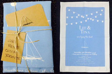 Custom printed wedding tea towel invitation in organic cotton with nautical blue design, folded with twine and tag, featuring ‘Lee & Tina are tying the knot’ print.