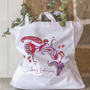 White organic cotton tote bag with a vibrant red, purple, and olive green paisley-style design and the custom text ‘Sam’s Foodie Goody’. Filled with eucalyptus branches, leaning against a rustic stone wall.