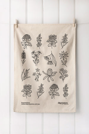 Natural tea towel with black native floral design hanging on a white wooden panel wall.