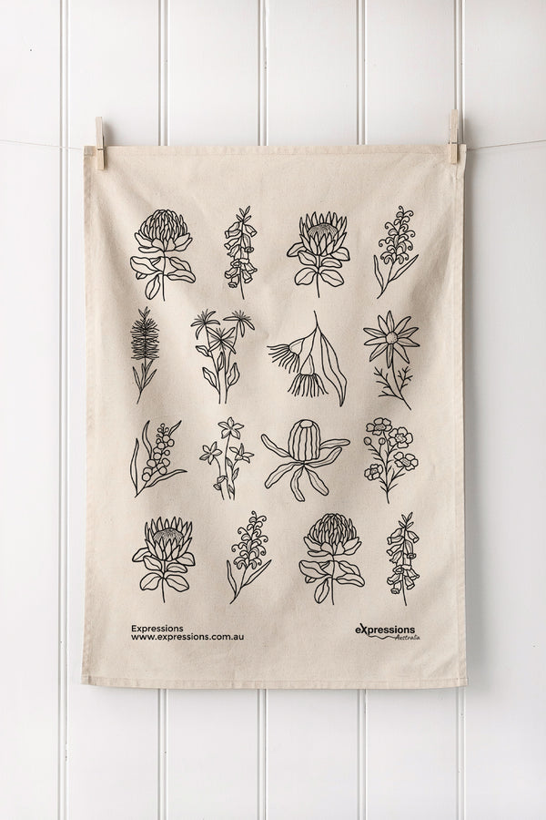Natural tea towel with black native floral design hanging on a white wooden panel wall.