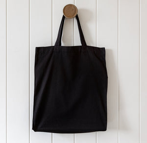 Black tote bag hanging on a wooden hook against a white wooden panel background