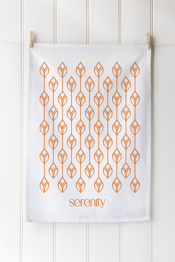 White tea towel with orange geometric pattern and logo at bottom on a white wooden background