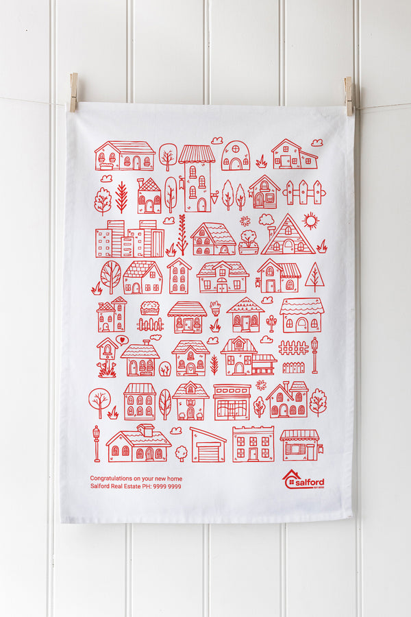White tea towel with red house pattern hanging on a wooden wall with customer logo and text.