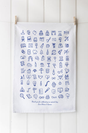 White tea towel with blue wedding icons hanging on a wooden rack against a white wall.